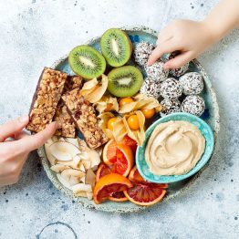 Mother sharing healthy vegan dessert snacks with toddler child. Concept of healthy sweets for children. Protein granola bars, homemade raw energy balls, cashew butter, toasted coconut chips, fruits platter